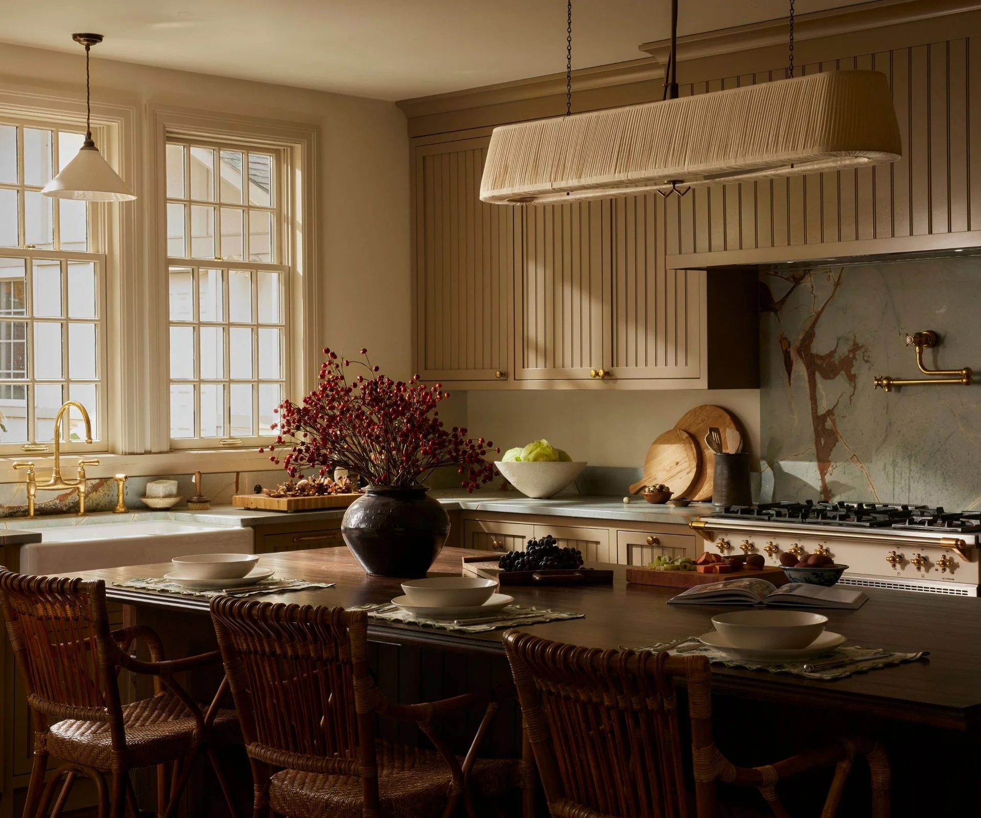 Cozy kitchen, large dark wood dining table in center, decorated with vase of flowers and ableware, slatted wood kitchen cabinets, stove and windows, sink in background