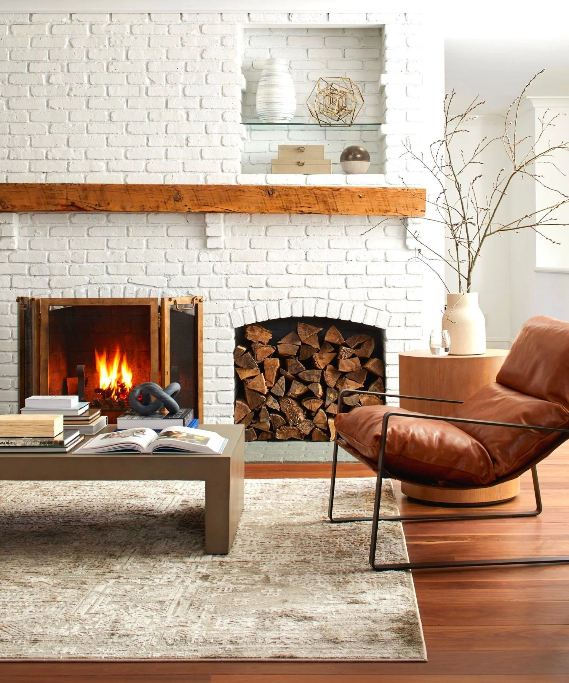 A living room with a white brick wall, a fireplace with logs next to it, a wooden coffee table, and a brown leather armchair