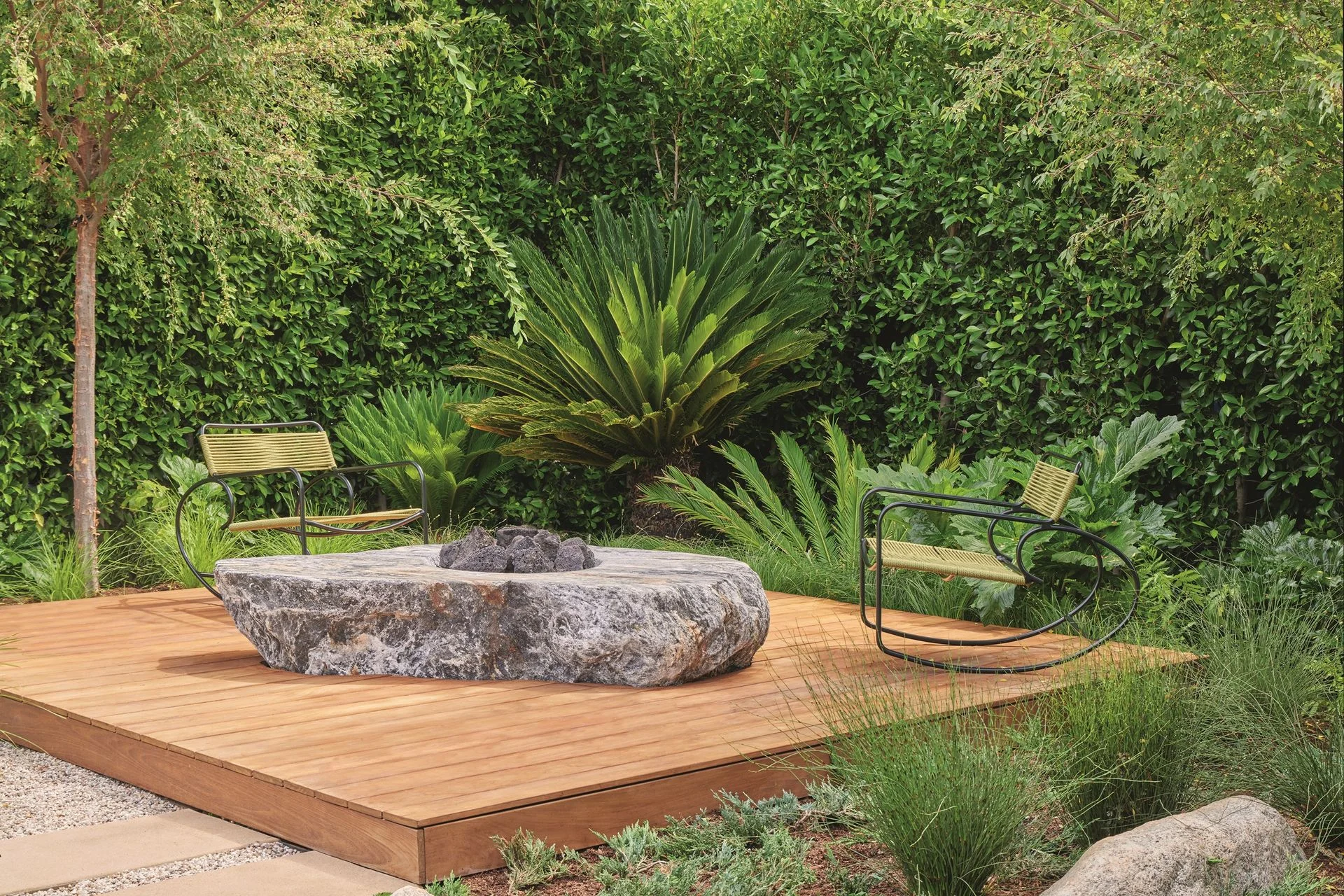 A wooden deck in a lush garden with a large stone fireplace in the center
