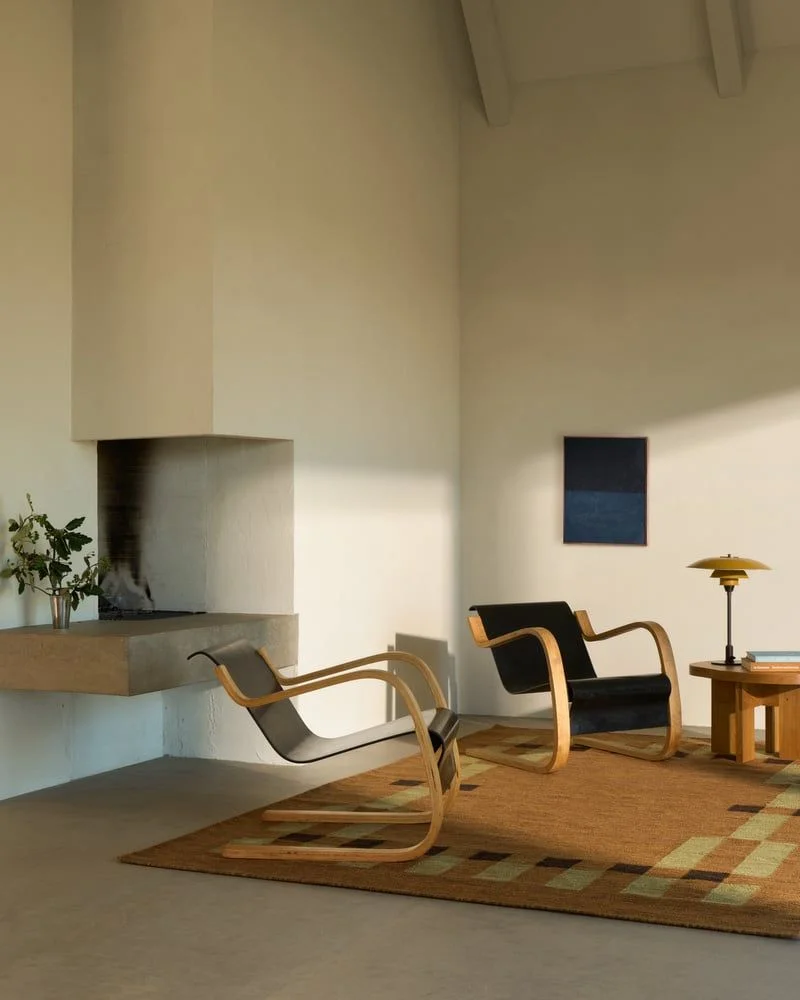 Image of a Scandinavian-style living room with white walls and a concrete floor. There is a tan flatweave rug with two modern accent chairs on it and a wooden side table.