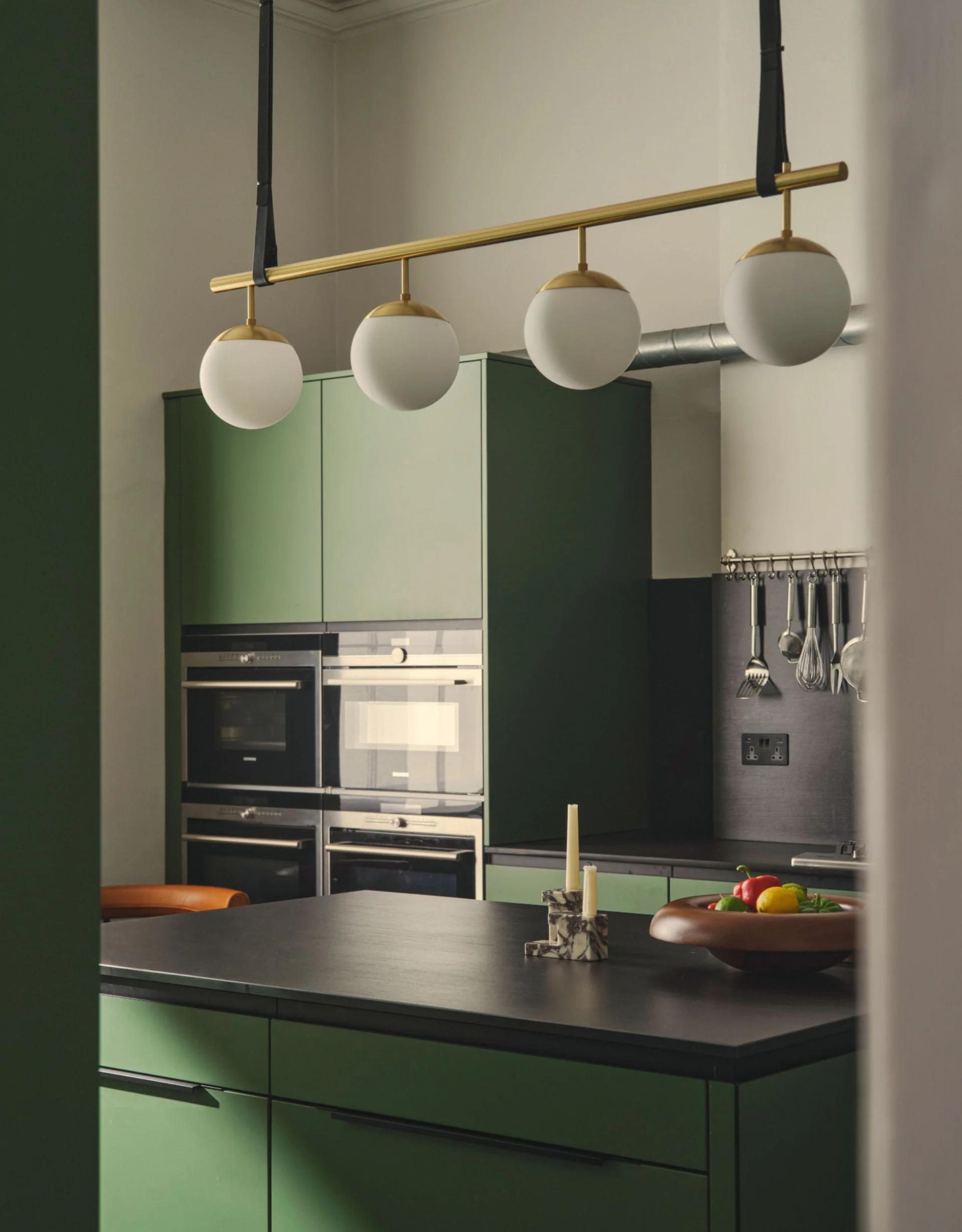Green modern kitchen with black countertop and splashback, and gold and white opal linear pendant light over the island
