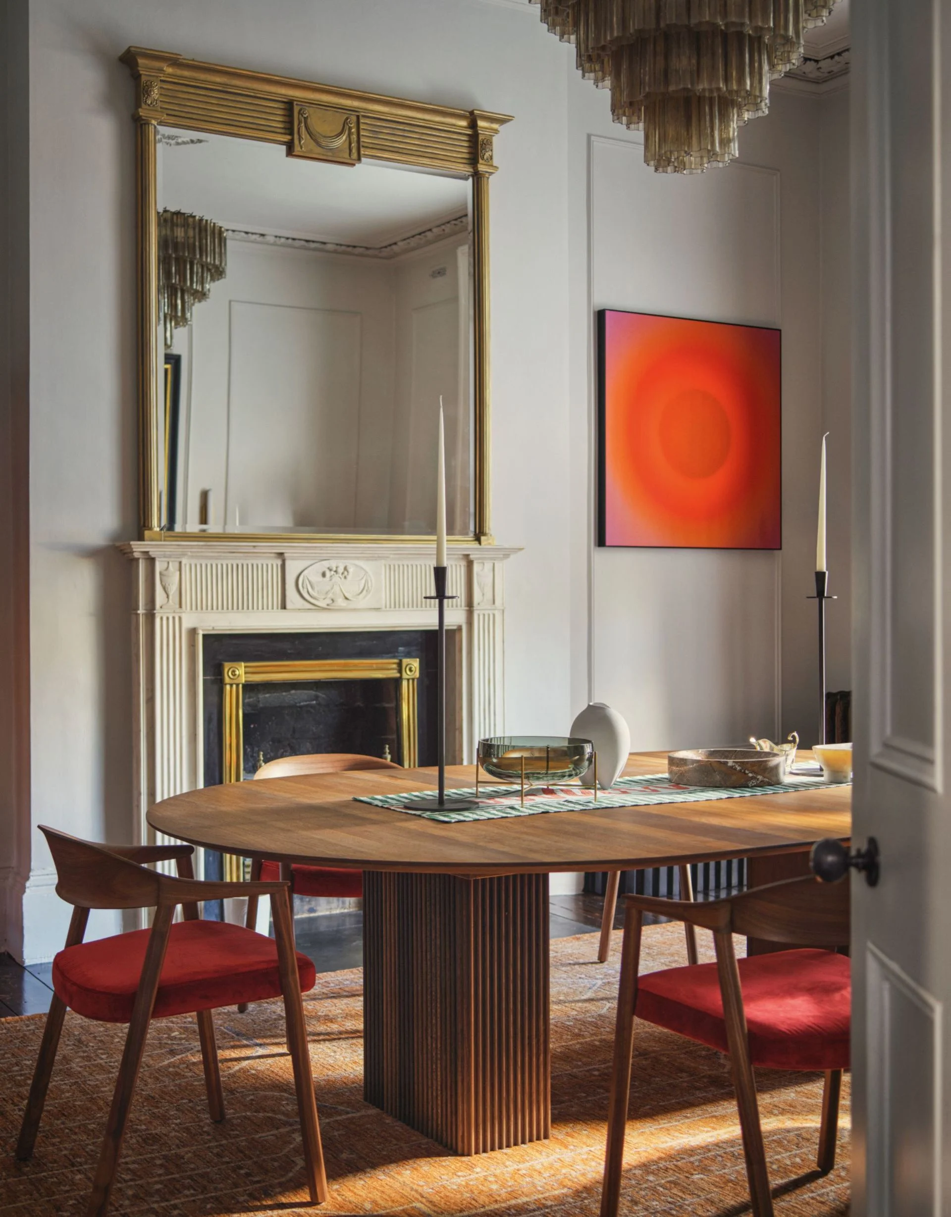 Dining room with pale grey walls, wood oval dining table with reeded legs, wood curved-back armchairs with red velvet seats, and abstract red-orange artwork