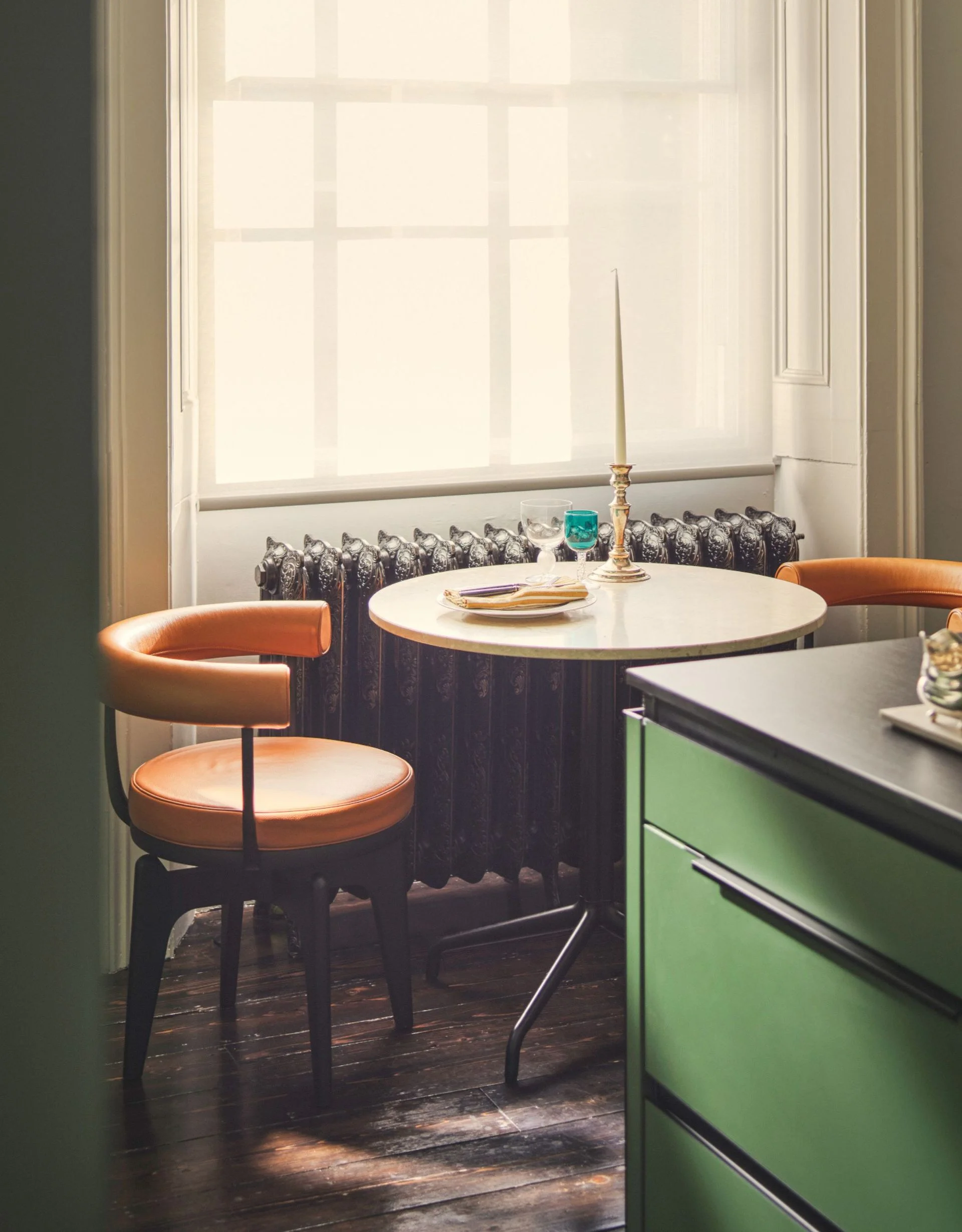 Corner of a green kitchen with a marble and black metal cafe table and orange leather curved-back dining armchairs