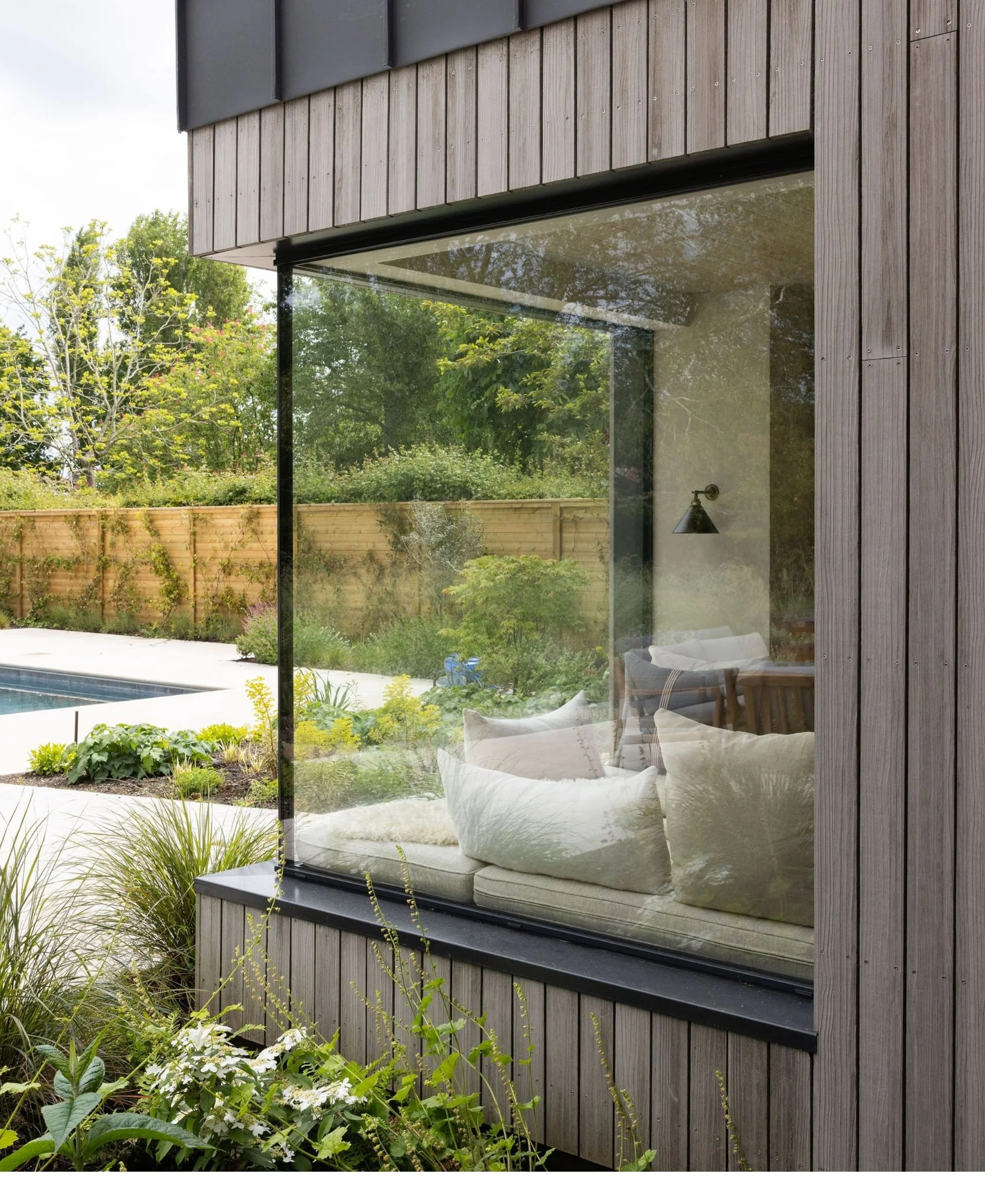 Frameless glass corner window overlooking a garden and pool, framing a cushioned window seat inside.
