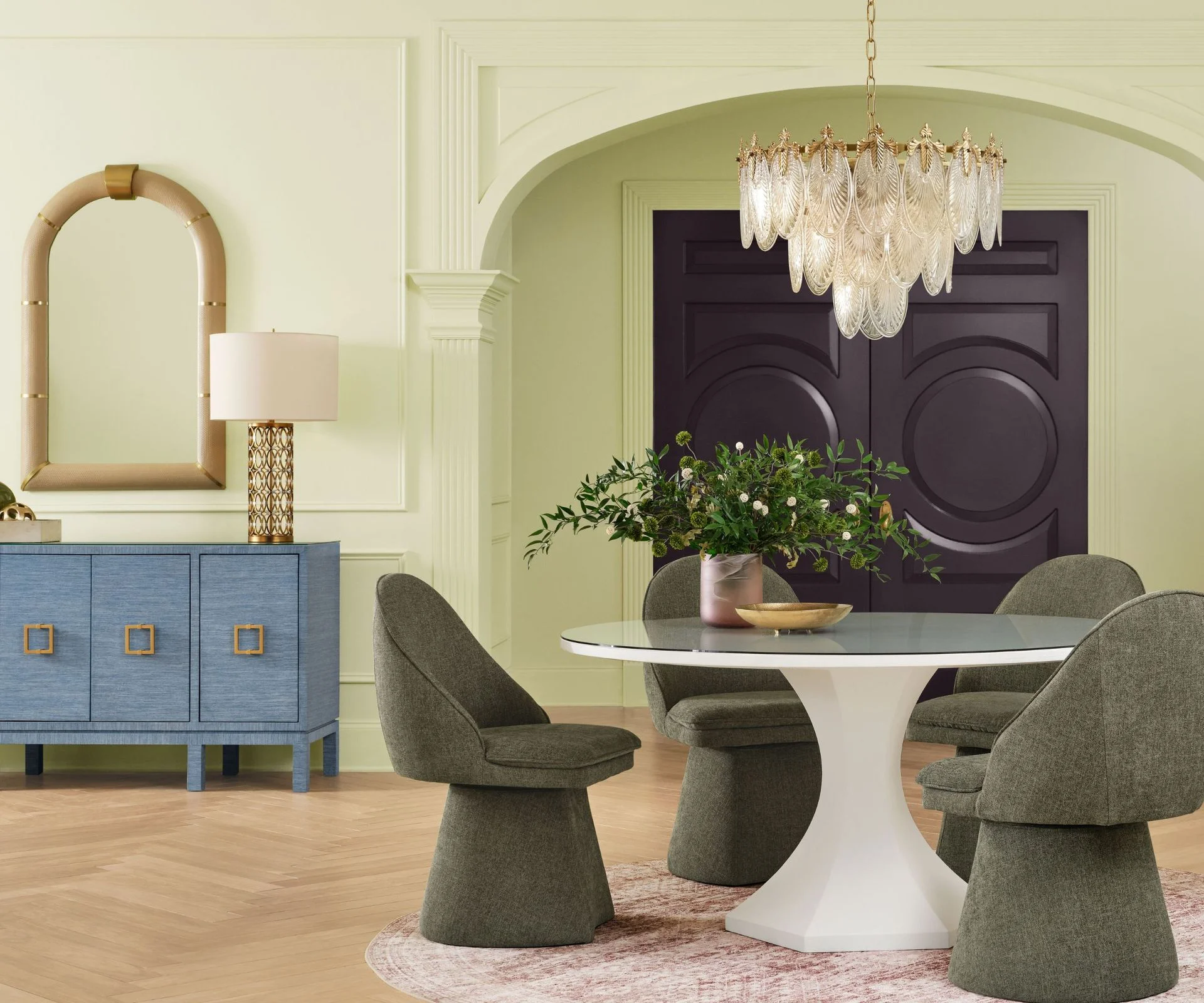 Dining room painted in Sherwin-Williams Celery, a dark aubergine front door, a round white table with dark green armchairs, and a blue console with a mirror and lamp.